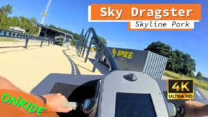 Blick aus der Onride-Perspektive des Sky Dragster im Skyline Park. Die Hände des Fahrers halten den Steuergriff des Motorrads, das auf einer schwarzen Schiene entlang einer sandfarbenen Strecke fährt. Im Hintergrund ist ein Gebäude mit der Aufschrift "Spike Racing" zu sehen. Die Aufnahme ist in 4K Ultra HD-Qualität.