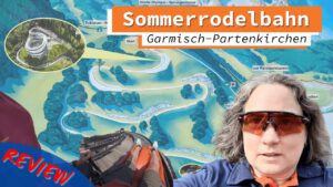 Person mit Sonnenbrille vor einer Karte der Sommerrodelbahn in Garmisch-Partenkirchen. Im Hintergrund Streckenverlauf der Rodelbahn und ein Detailbild der Strecke. Eine Rodelperspektive und der Schriftzug „Review“ ergänzen das Bild.