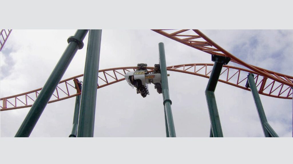 Die Achterbahn Sierra Sidewinder mit drehbaren Wagen auf einer orangefarbenen Strecke zwischen grünen Stützen und wolkigem Himmel.