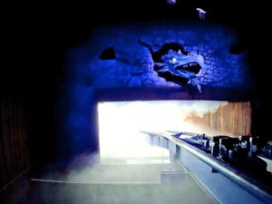 Innenaufnahme der Achterbahn Fenix im Toverland, mit einer thematischen Drachenprojektion an einer Steinwand und einer Schiene, die in einen Nebelbereich führt.