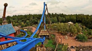 Onride-Perspektive der Fenix-Achterbahn im Toverland mit blauen Schienen, einer steilen Abfahrt und thematisch gestalteter Umgebung aus Gebäuden und Wald.