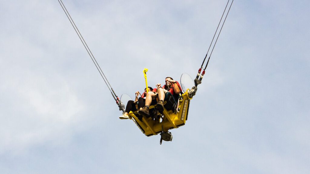 Fahrgäste in einer gelben Gondel des Funtime Slingshot, die an zwei starken Seilen befestigt ist und hoch in den Himmel geschleudert wird.