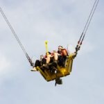 Fahrgäste in einer gelben Gondel des Funtime Slingshot, die an zwei starken Seilen befestigt ist und hoch in den Himmel geschleudert wird.
