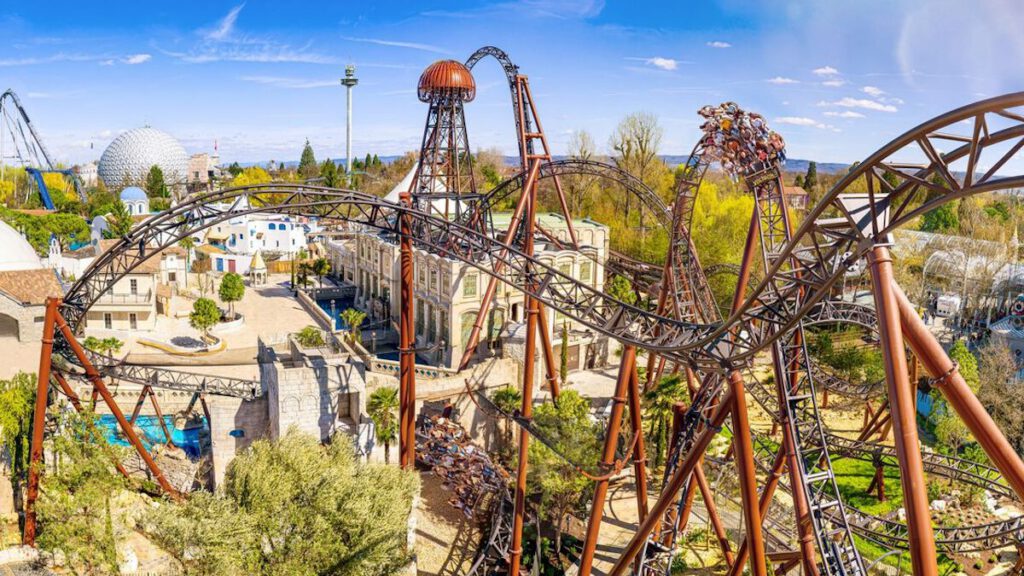 Der Stryker Coaster mit verschlungenen braunen Schienen und mehreren Inversionen, eingebettet in eine thematische Umgebung mit mediterranen Gebäuden und grüner Landschaft.