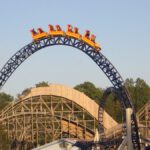 Achterbahnzug auf einem Top Hat-Element mit steilem Auf- und Abstieg, gelben Wagen und einer blauen Schiene, umgeben von Holzkonstruktionen und Bäumen.