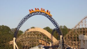 Achterbahnzug auf einem Top Hat-Element mit steilem Auf- und Abstieg, gelben Wagen und einer blauen Schiene, umgeben von Holzkonstruktionen und Bäumen.