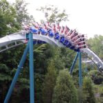 Achterbahnzug in einer Zero-G-Roll, bei der die Schiene eine Schraubenbewegung ausführt, während Fahrgäste kopfüber durch die Strecke gleiten, umgeben von Bäumen.