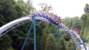 Achterbahnzug in einer Zero-G-Roll, bei der die Schiene eine Schraubenbewegung ausführt, während Fahrgäste kopfüber durch die Strecke gleiten, umgeben von Bäumen.