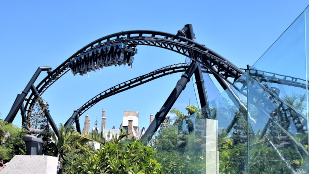 Ein Zero-G-Stall auf einer Achterbahn mit schwarzer Schiene, bei dem der Zug kopfüber verharrt, umgeben von grüner Vegetation und thematisierten Gebäuden.