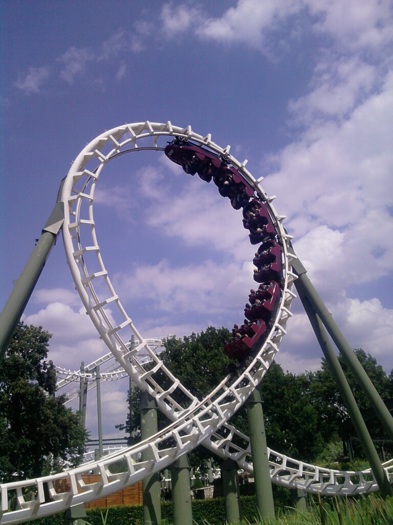 Eine Achterbahn mit weißer Schiene und grünen Stützen durchfährt einen vertikalen Looping. Der Zug mit dunkelroten Wagen befindet sich kopfüber am höchsten Punkt der Inversion. Im Hintergrund sind weitere Schienen und Bäume unter einem leicht bewölkten Himmel zu sehen.