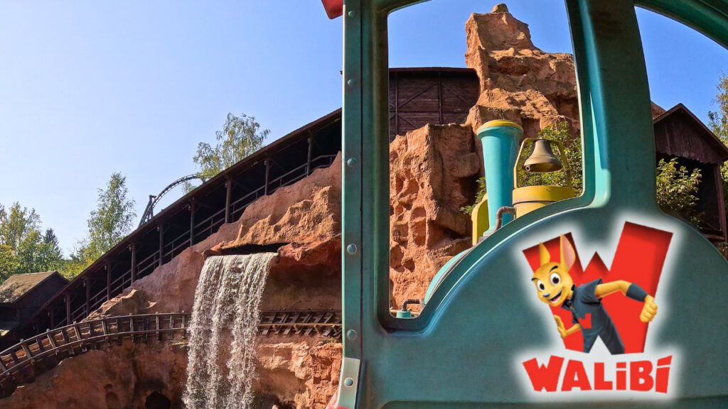Ein türkisfarbener Westernzug fährt durch eine felsige Themenlandschaft mit einem künstlichen Wasserfall im Freizeitpark Walibi. Im Hintergrund ist eine hölzerne Achterbahnstruktur sichtbar, umgeben von Bäumen. Rechts unten ist das Walibi-Logo mit einem Comic-Maskottchen zu sehen.